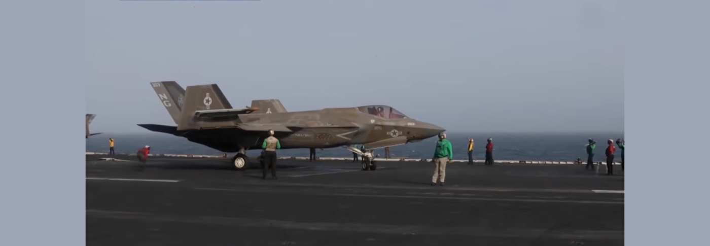 An F-35 fighter jet sits on an aircraft carrier flight deck at sea, surrounded by Navy deck crew in colored jerseys.